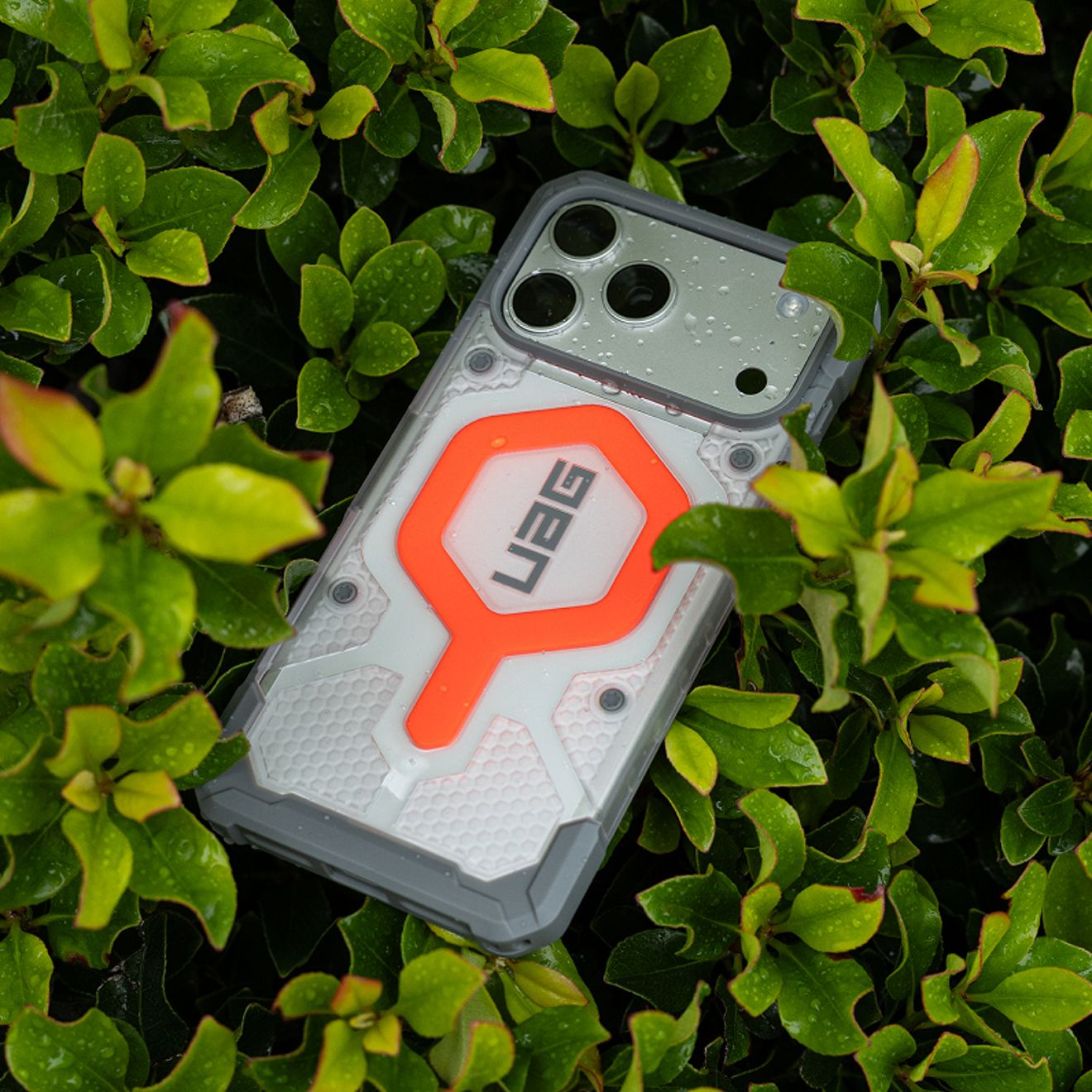 Un teléfono con una funda resistente UAG en tonos blancos y grises, con detalles en naranja brillante, descansa entre hojas verdes húmedas, con gotas de agua visibles tanto en el teléfono como en la funda.