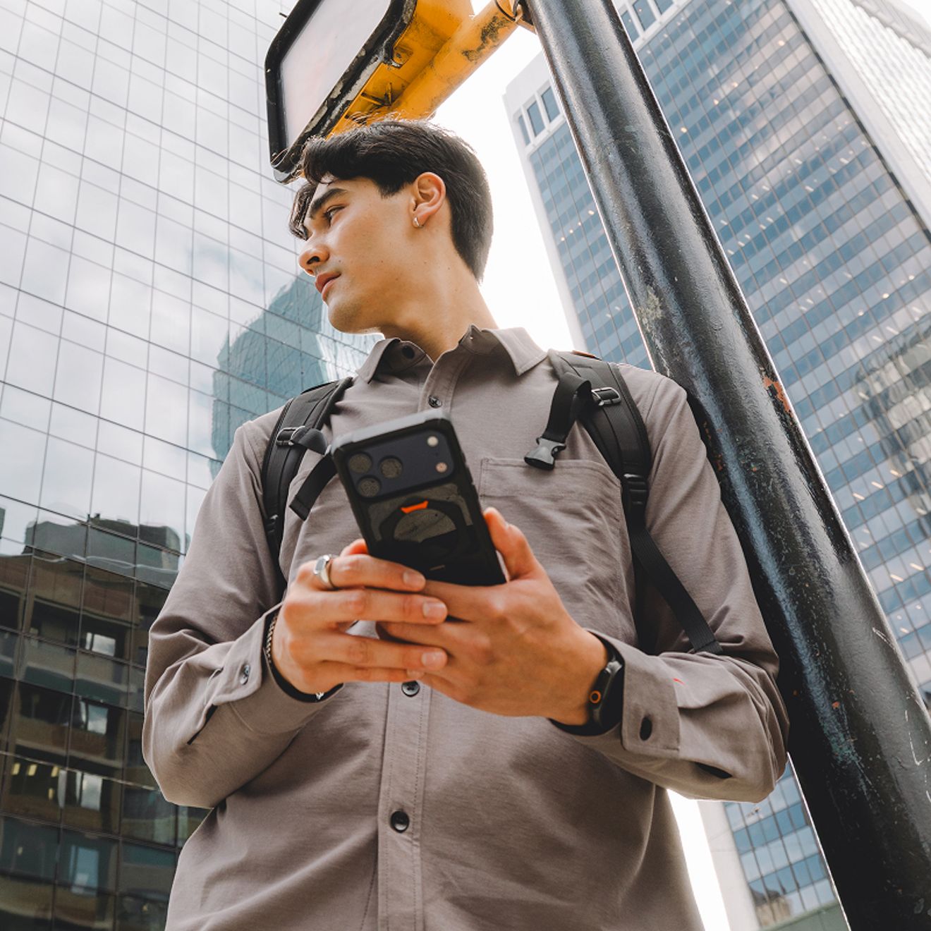 Una persona con mochila sostiene un smartphone protegido por una funda resistente UAG mientras se encuentra entre edificios altos en una ciudad.