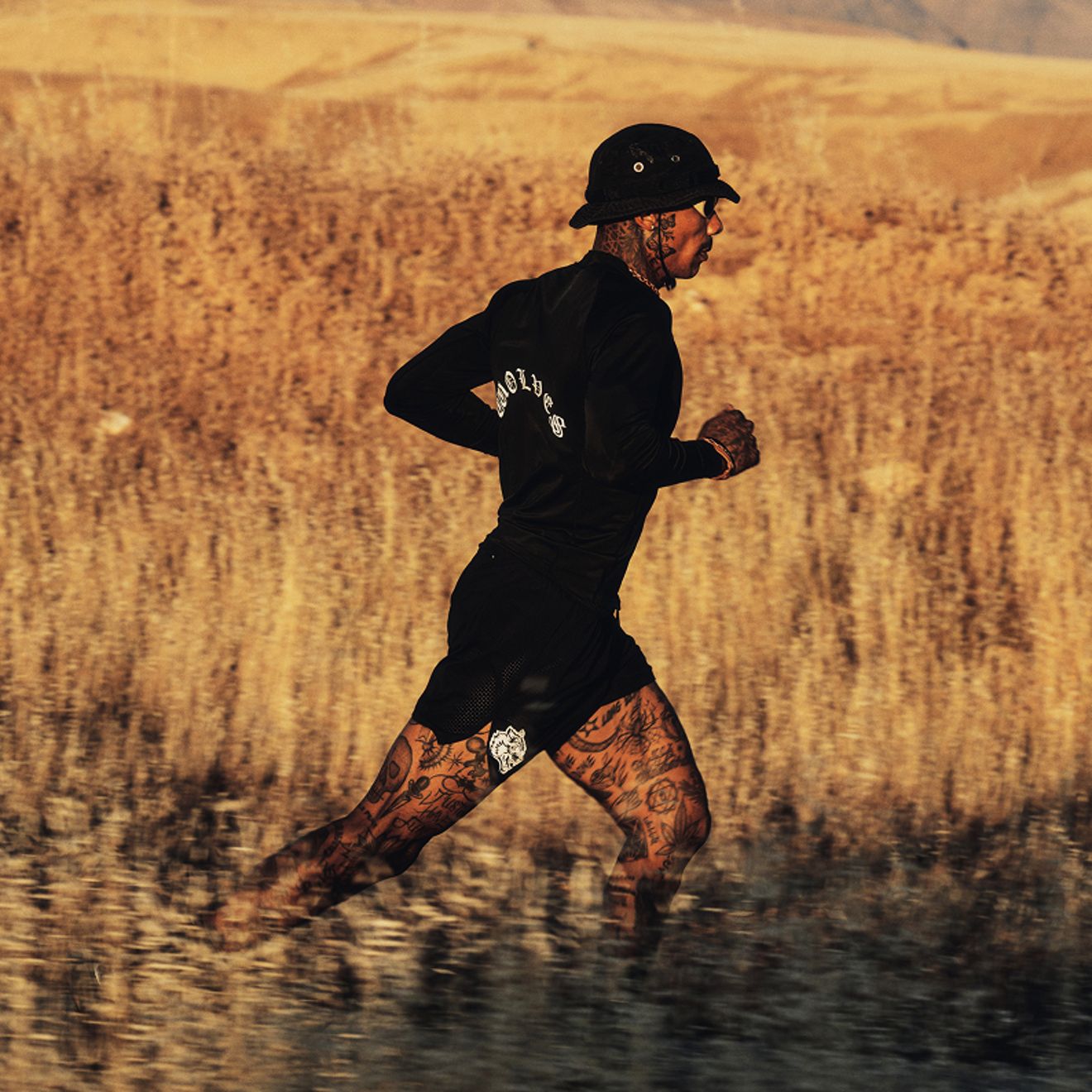 Un corredor tatuado, vestido con ropa deportiva negra, un sombrero de pescador y gafas de sol, corre por un campo bañado por la luz del sol.