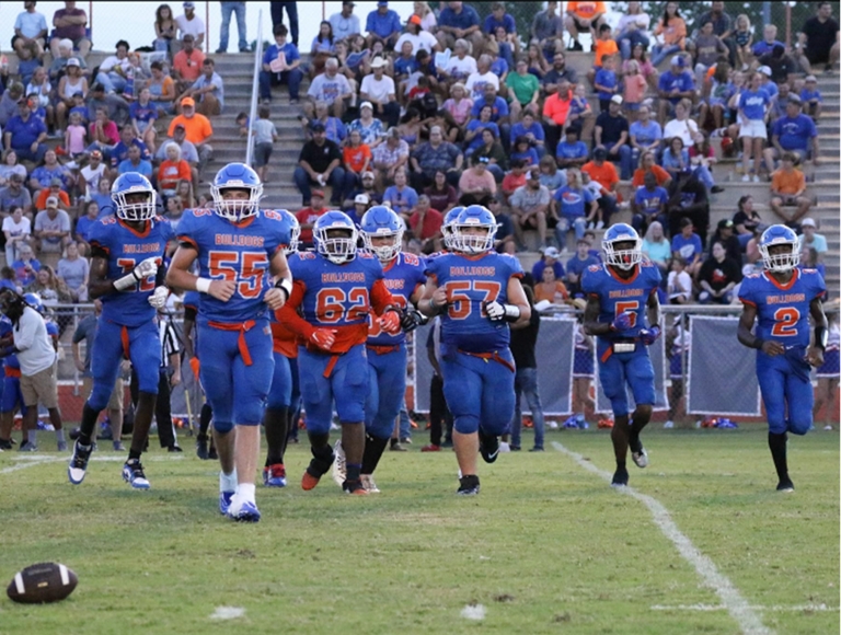 El equipo de fútbol de la Taylor County High School, vestido con uniformes azules y naranjas, corre hacia el campo.