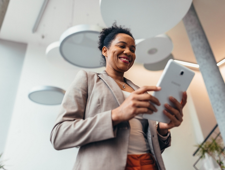 Una persona sonríe y mira su tablet digital mientras está de pie en el vestíbulo de un edificio moderno.