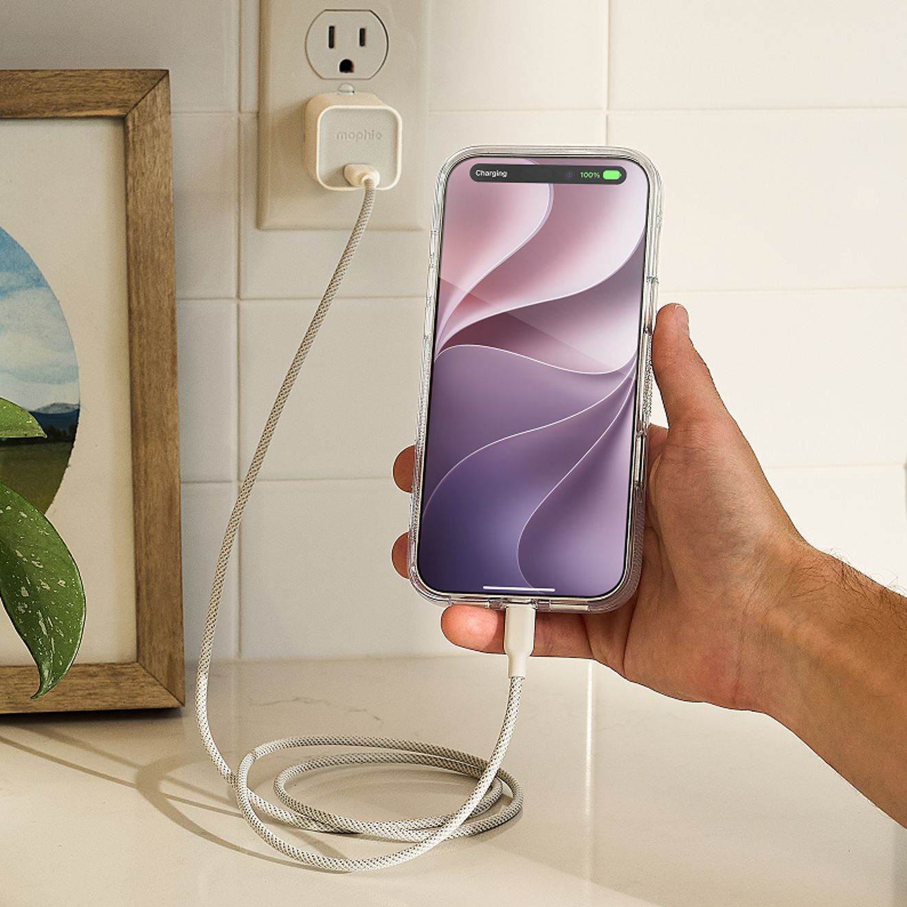 Una mano sostiene un iPhone color plata con una funda transparente mientras se carga mediante un cable USB-C trenzado blanco conectado a un cargador de pared Mophie USB-C de 35 W blanco, enchufado a un tomacorriente.