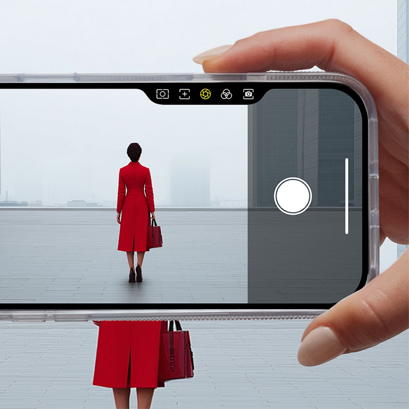 Una mano sostiene un iPhone en posición horizontal dentro de una funda transparente y utiliza el botón integrado de la cámara para encuadrar la foto de una mujer con abrigo rojo, situada frente a un edificio moderno.