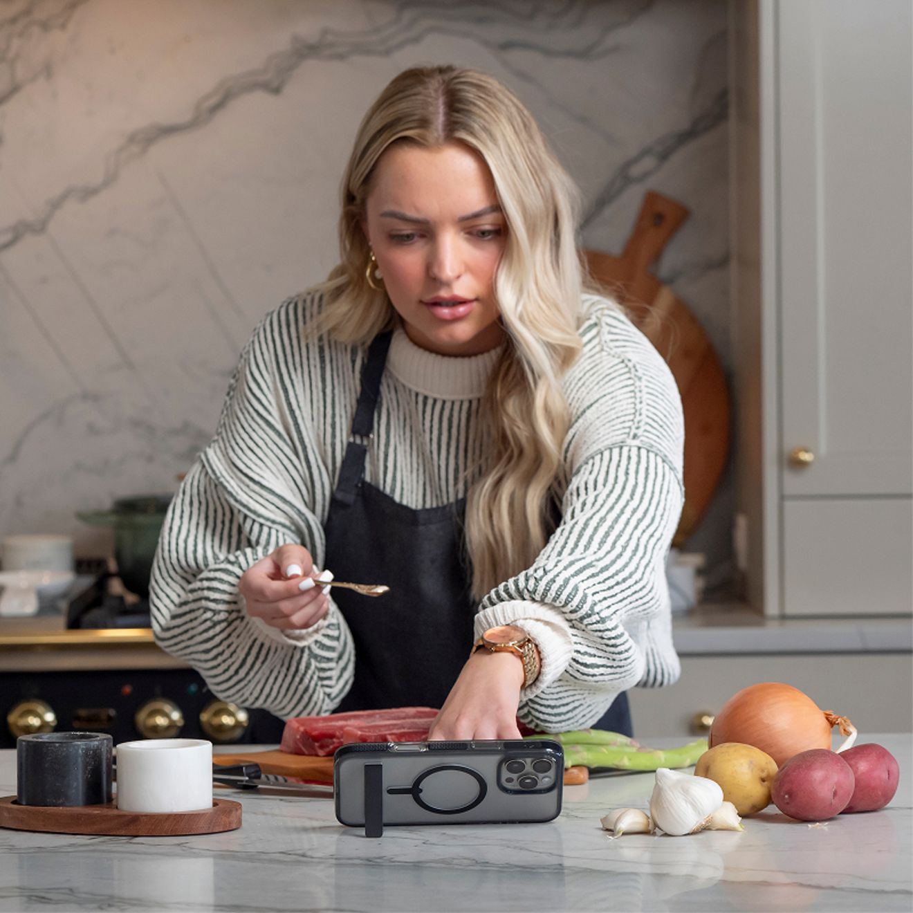 Una mujer prepara comida en una cocina mientras un iPhone Pro plateado, dentro de una funda transparente ZAGG con anillo magnético y base de apoyo integrada, está apoyado horizontalmente sobre la encimera.
