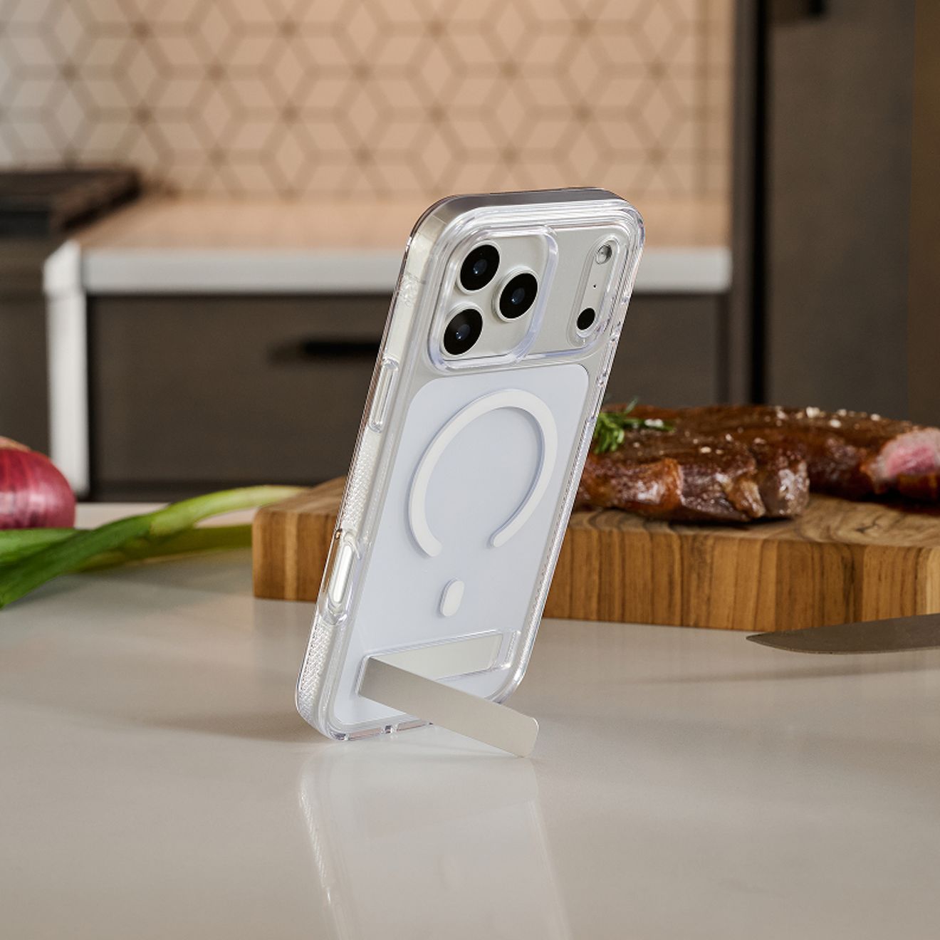 Un iPhone 17 Pro plateado, dentro de una funda transparente ZAGG Crystal Palace con un anillo de carga magnética blanco y una base de apoyo integrada, se mantiene en posición vertical sobre una encimera de cocina.