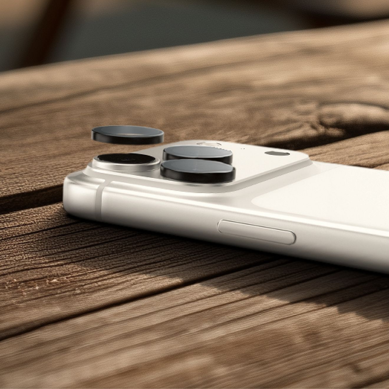 Primer plano de un iPhone Pro color plata sobre una superficie de madera, en el que se están retirando los protectores para lentes de cámara ZAGG Glass Elite de la cámara trasera con la herramienta de extracción incluida.