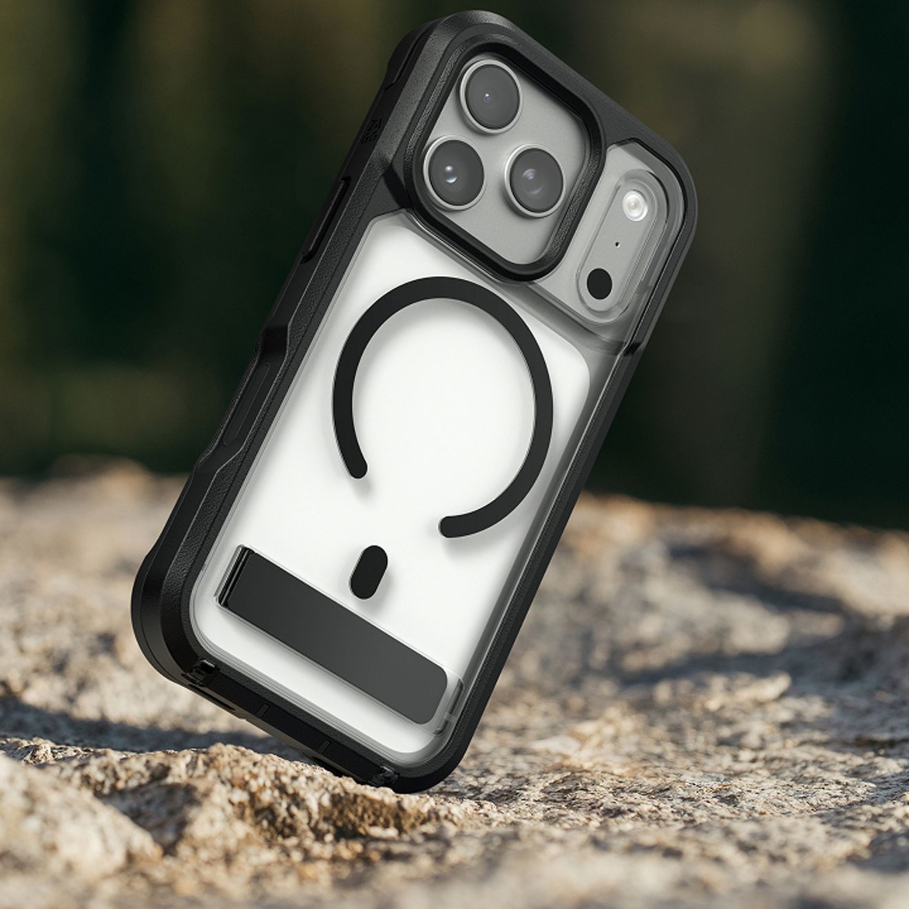 A silver iPhone Pro in a clear and black protective case with a magnetic charging ring and built-in kickstand stands upright on a rocky outdoor surface.