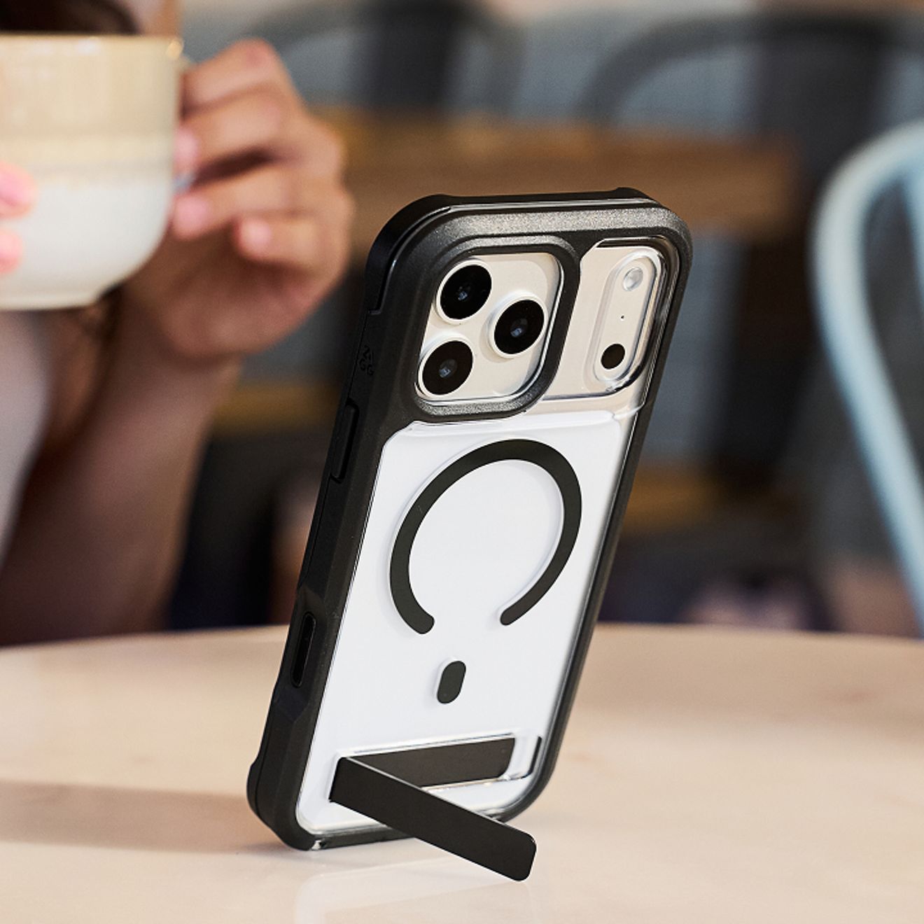 A silver iPhone 17 Pro in a clear and black protective case with a magnetic charging ring is propped upright on a tabletop using its built-in black kickstand.