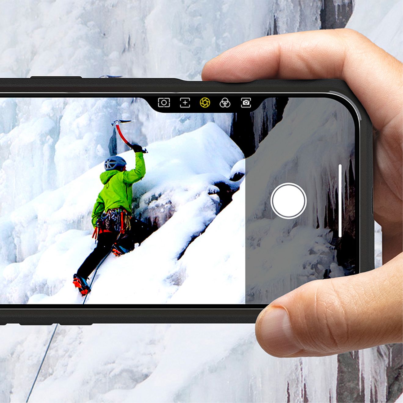 A hand holds an iPhone horizontally in a black protective case, using the integrated camera button to frame a photo of an ice climber scaling a snowy frozen cliff.