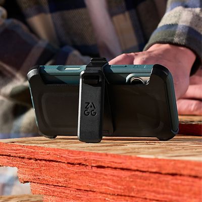 Primer plano de un iPhone 17 Pro plateado en una funda protectora ZAGG Teton azul, sujeta a un portateléfono ZAGG negro con clip para cinturón, apoyado sobre una superficie de madera.