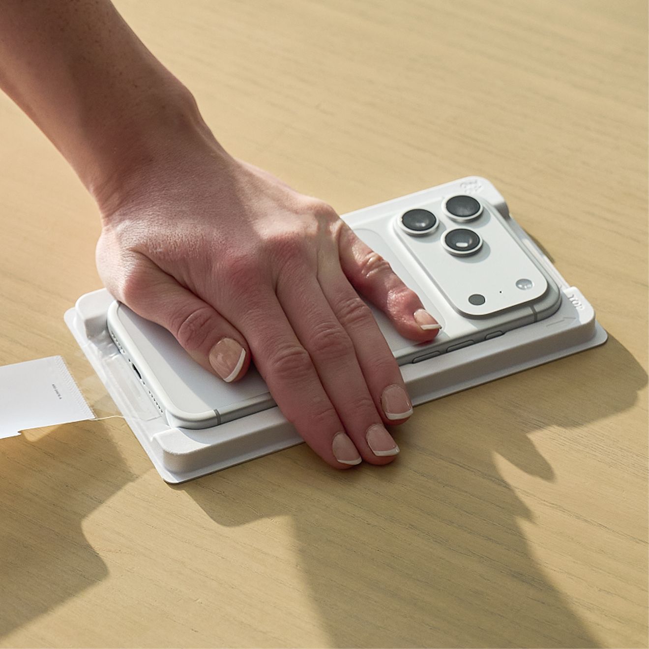 Una mano presiona un iPhone 17 Pro plateado, fijado en una bandeja de instalación ZAGG PerfectFit™ blanca sobre una mesa de madera, para mostrar cómo se coloca el protector de pantalla.