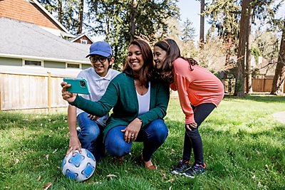 Una mujer y dos niños agachados junto a un balón de fútbol en un jardín para tomar un selfie.