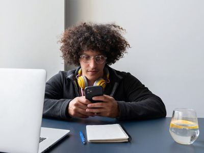 Hombre joven con audífonos alrededor del cuello y un teléfono en la mano sentado a una mesa con una computadora, un anotador y una bebida.