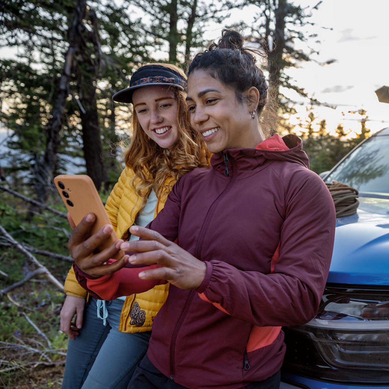 Dos mujeres están de pie junto a un SUV en un bosque, sonriendo mientras miran juntas un smartphone.