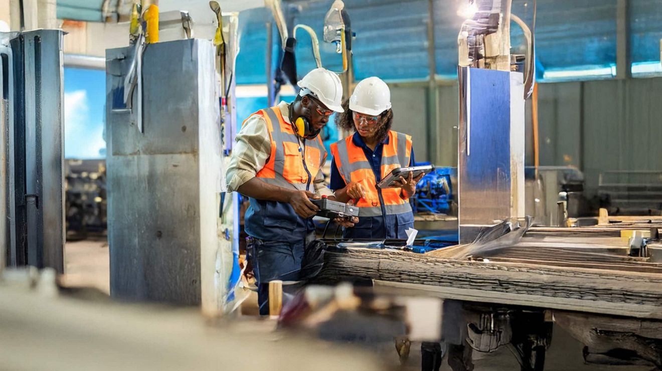 Dos ingenieros mecánicos con casco utilizando equipos conectados de T-Mobile para solucionar problemas con maquinaria industrial.
