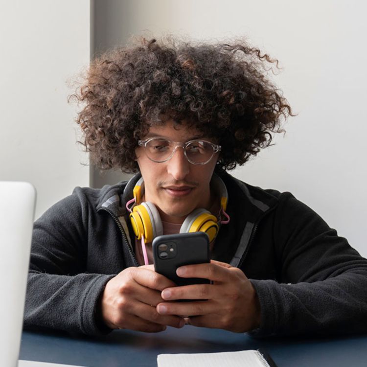 Hombre joven sentado en un escritorio con auriculares colgando del cuello y observando su teléfono.