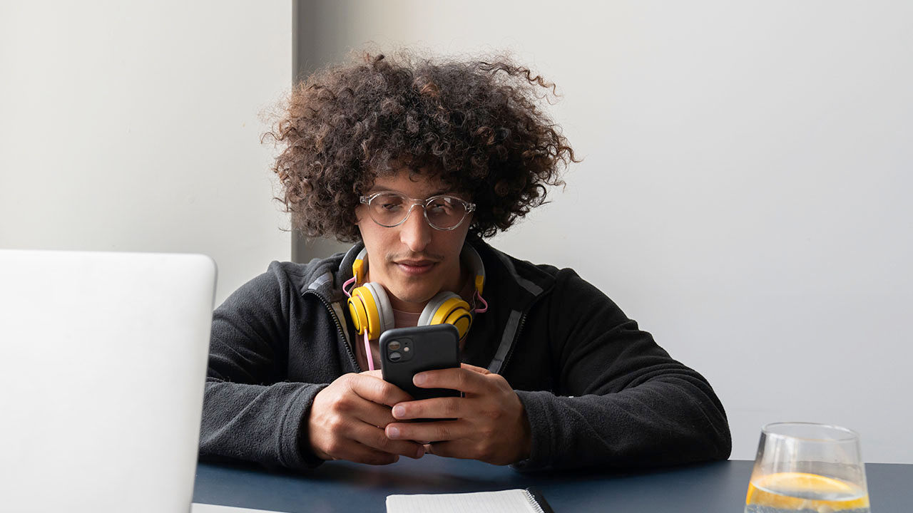 Hombre joven sentado a un escritorio con audífonos colgados al cuello y mirando un teléfono.