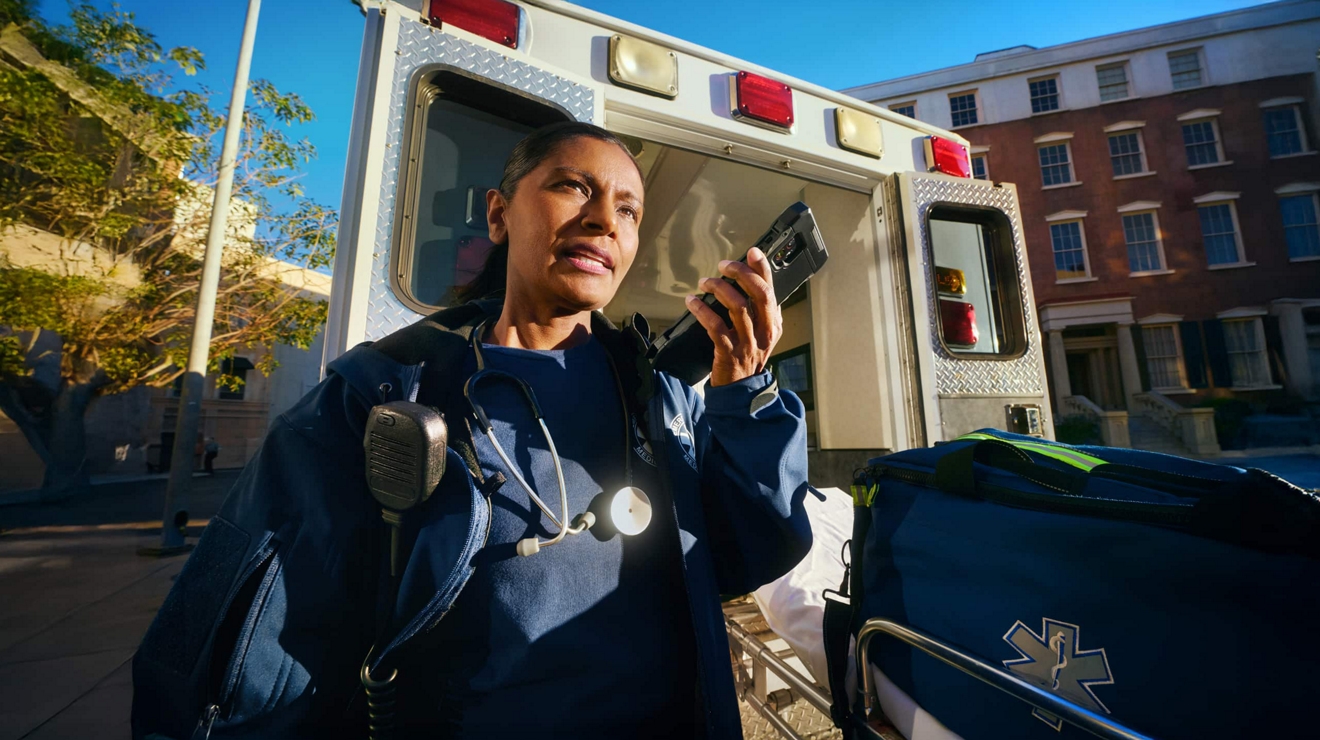 Miembro de servicios de emergencia comunicándose a través de un dispositivo MCPTT de pie delante de una ambulancia.