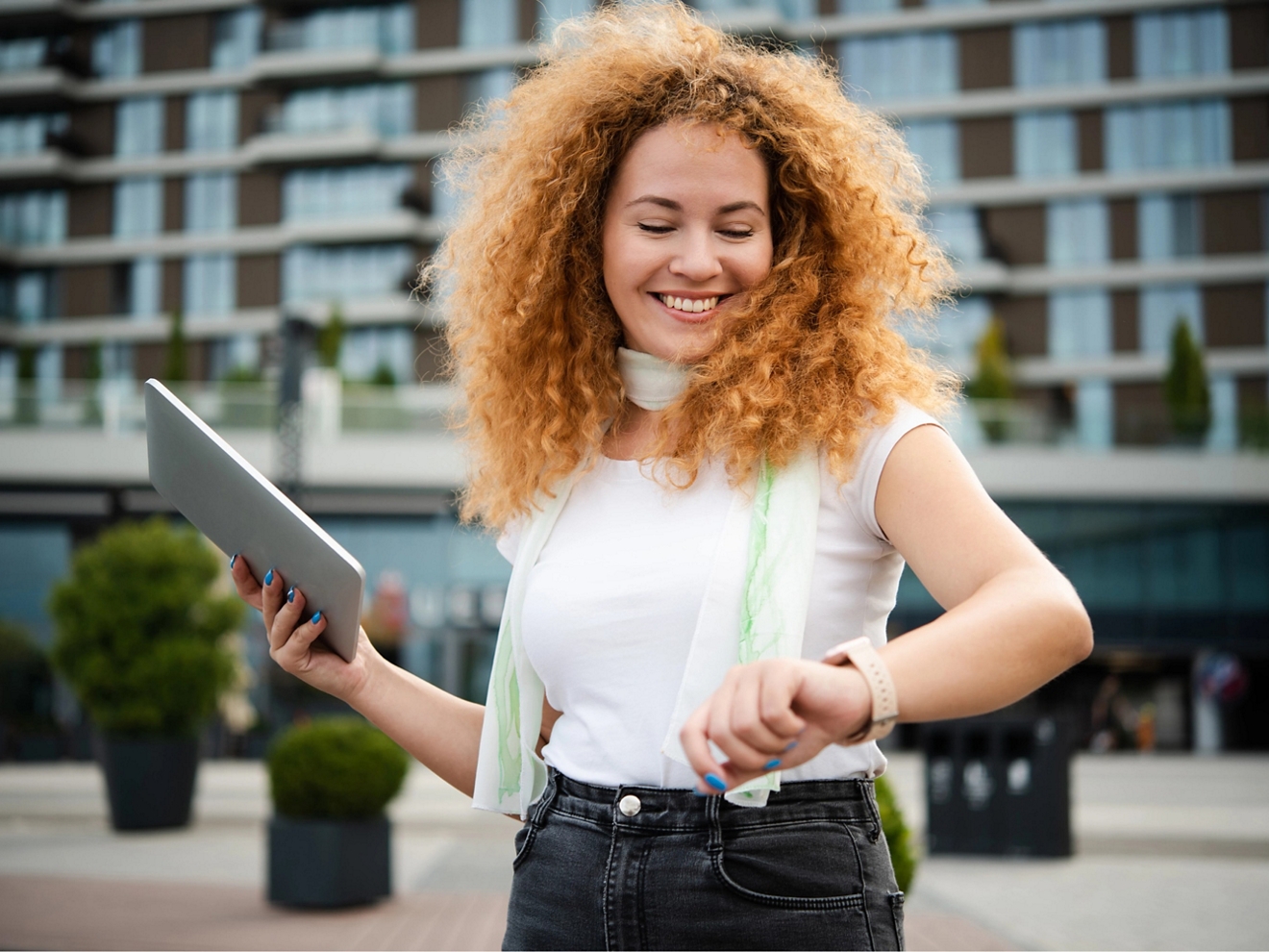 Persona sonriente en el exterior de un edificio de apartamentos, sosteniendo una tablet y consultando su reloj inteligente.