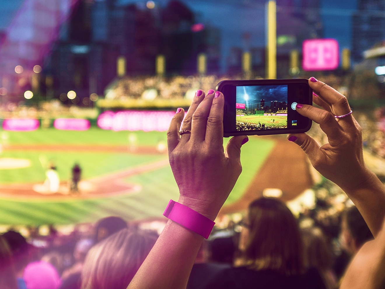 Las manos de una persona sosteniendo su teléfono mientras toma una foto del estadio de béisbol con la marca T-Mobile al fondo.
