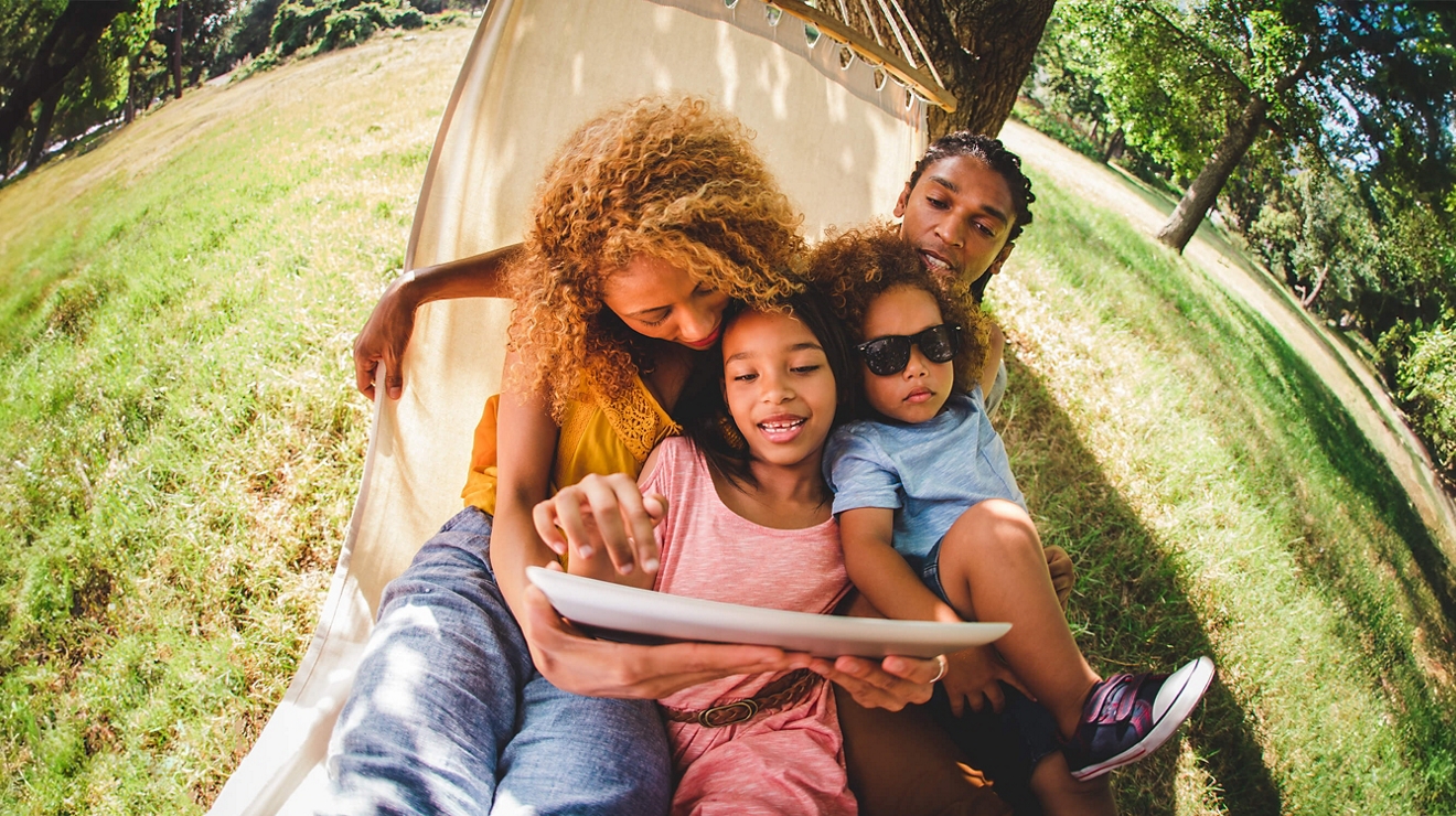 Una familia de cuatro sentada en una hamaca en un parque cubierto de césped, sonriendo a la pantalla de una tablet.