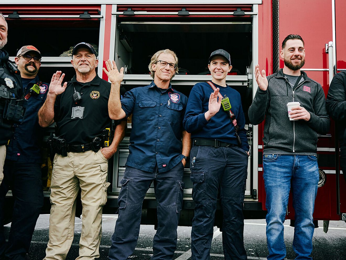 Un grupo de cinco bomberos y miembros de los servicios de emergencia están delante de un camión de bomberos rojo, sonriendo y saludando a la cámara.
