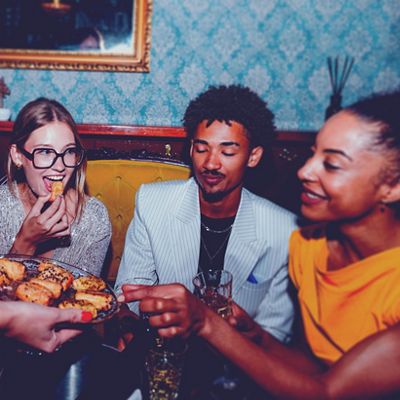 Tres amigos disfrutan de bocadillos y bebidas durante una animada reunión en interiores, sentados en un sofá de terciopelo con un papel tapiz de damasco azul de fondo.