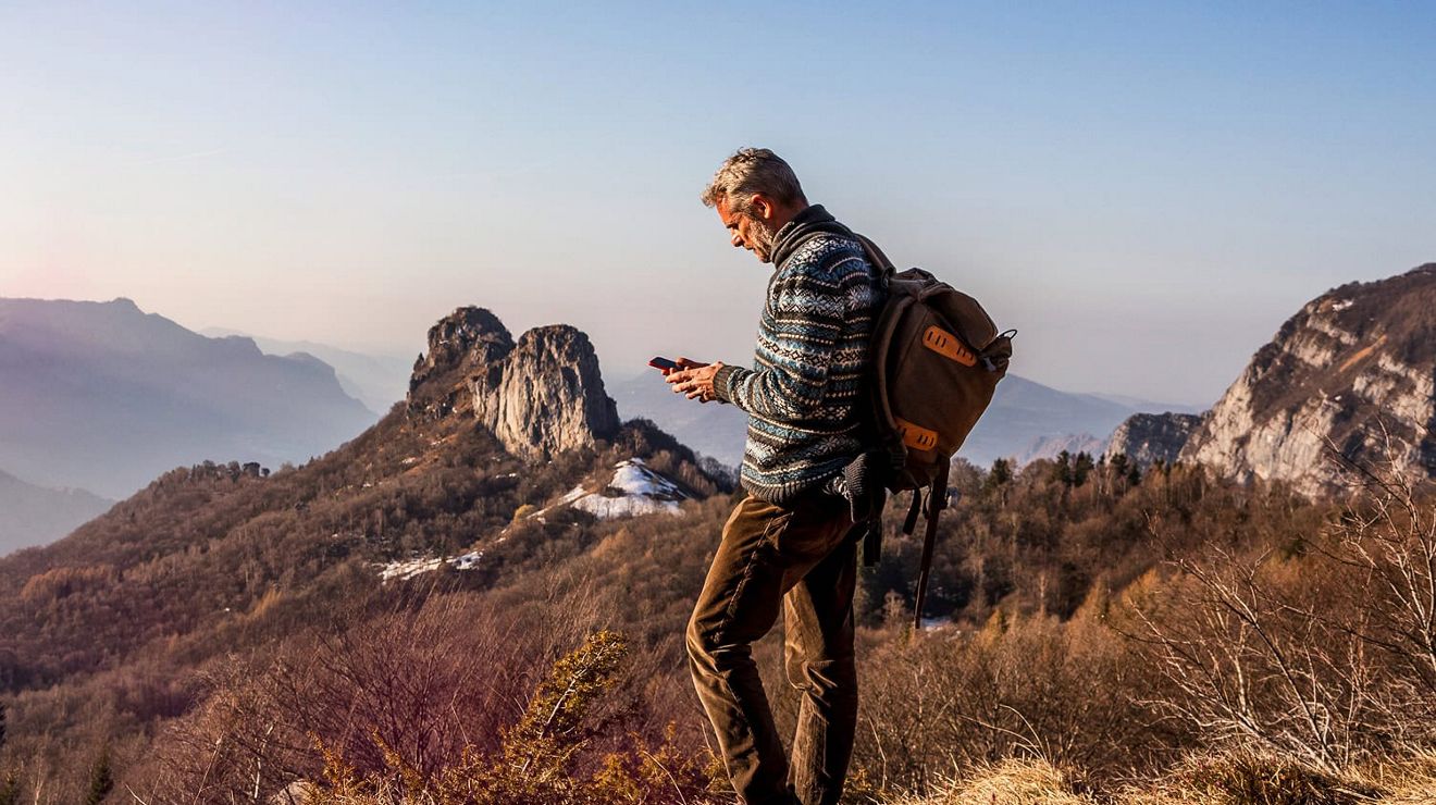 Un hombre con una mochila está de pie en una zona cubierta de hierba en lo alto de las montañas, hablando por teléfono.
