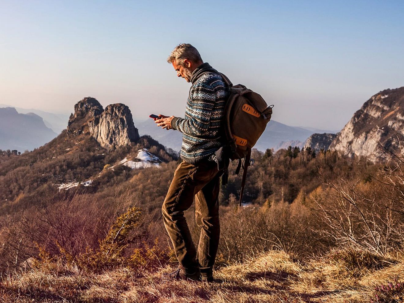 Un hombre con una mochila está de pie en una zona cubierta de hierba en lo alto de las montañas, hablando por teléfono.