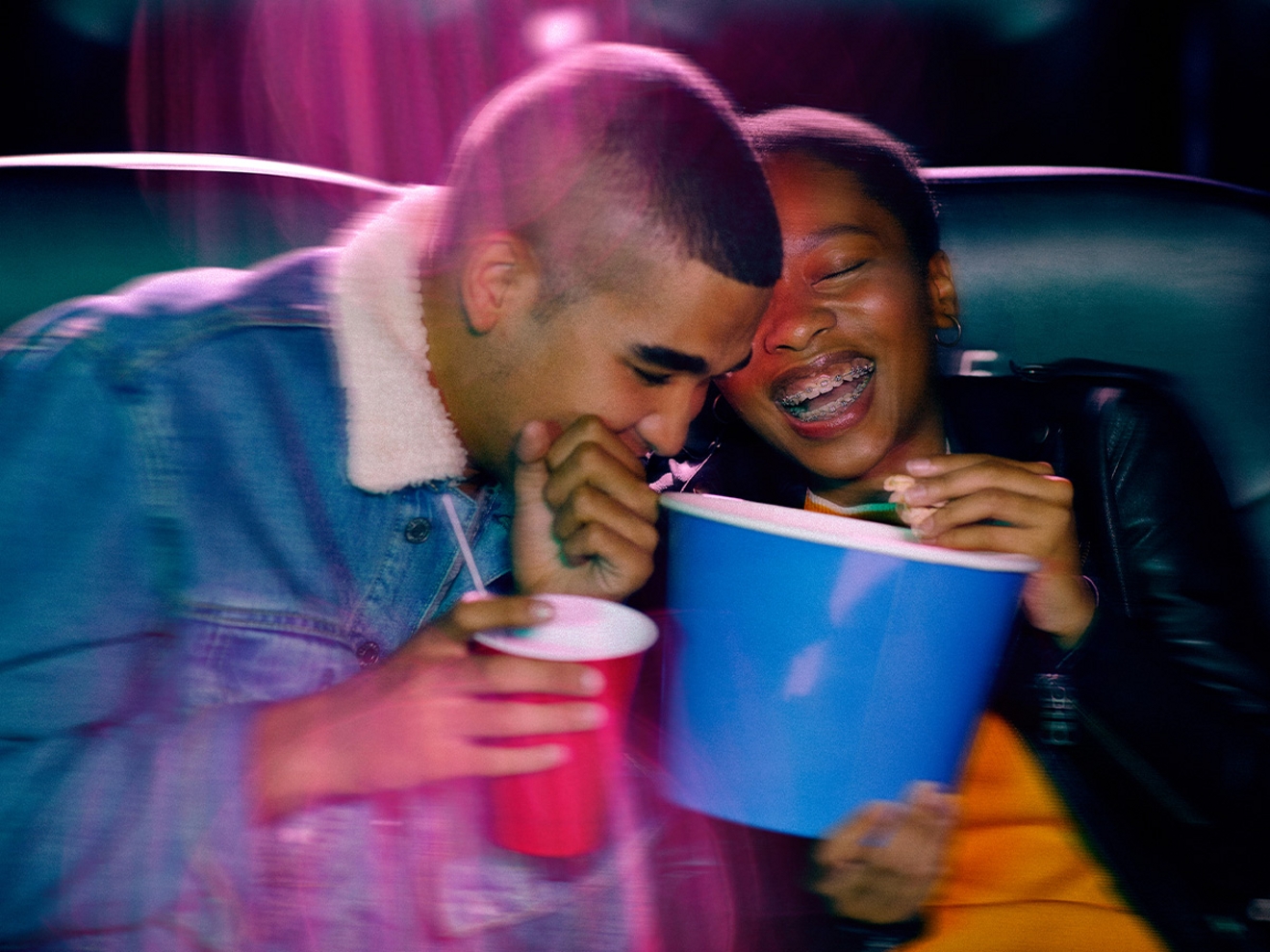 Dos amigos en un cine riéndose juntos en sus asientos, mientras uno sostiene un balde de palomitas y el otro un refresco.