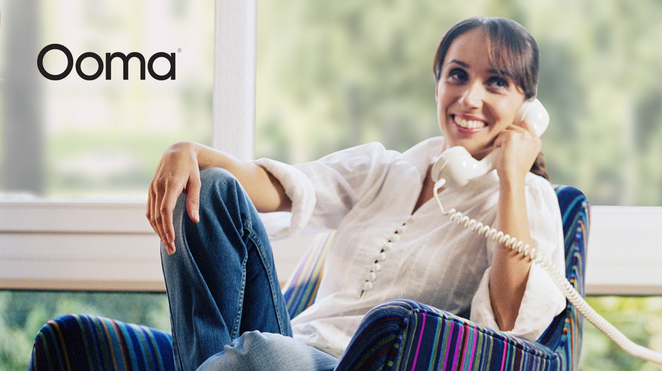 Ooma. Mujer sonríe y habla por teléfono de línea fija mientras está reclinada en una silla.