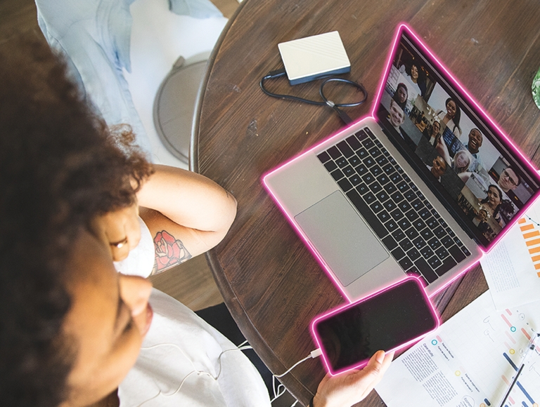 Toma aérea de una persona que sostiene un smartphone mientras trabaja en una laptop con papeles, una taza de café y cactus a su alrededor.