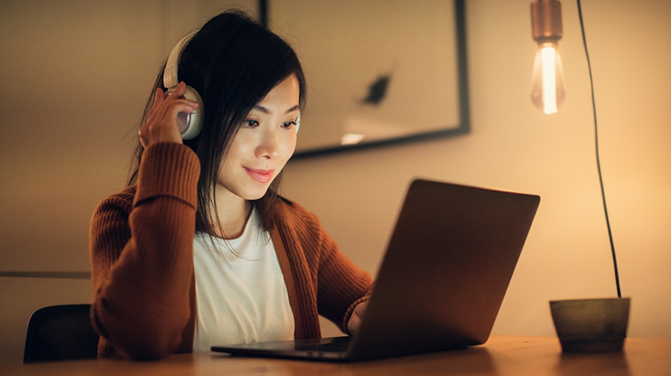 Una mujer sonríe sentada en un escritorio con audífonos puestos mientras mira la pantalla de su laptop.