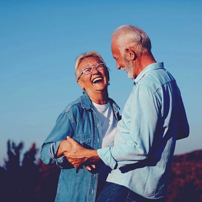 Una pareja de adultos mayores riendo al aire libre bajo un cielo azul despejado, tomados de la mano y mirándose el uno al otro.