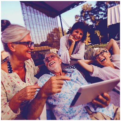 Un grupo de cuatro adultos mayores riendo y disfrutando de un momento juntos en un patio soleado, mientras una de las personas sostiene una tablet.