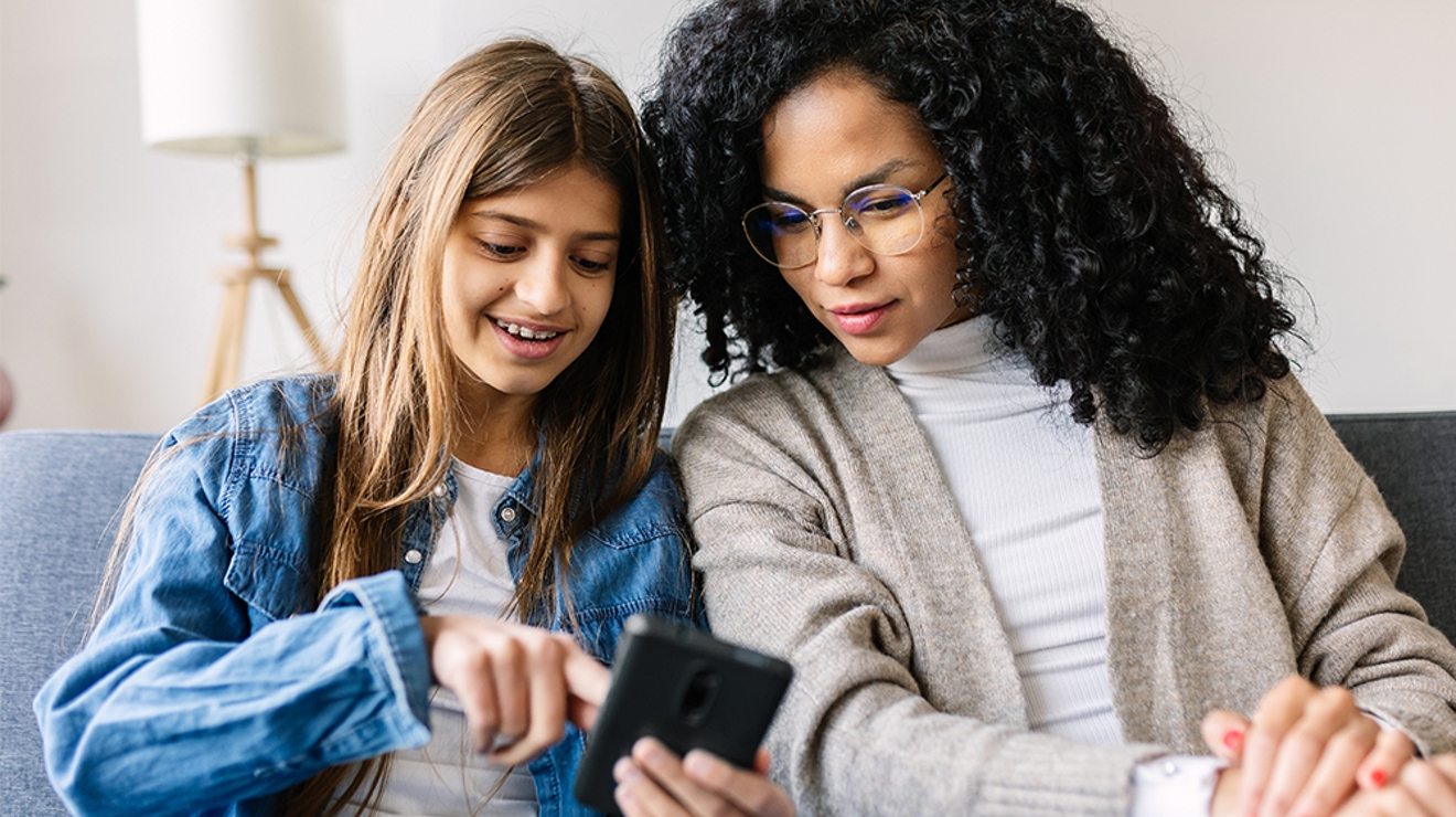 Una mujer y una niña sentadas en un sofá. Ambas sonríen mientras miran el teléfono de la niña.