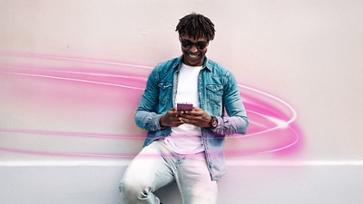 Un hombre con camiseta blanca, chaqueta de mezclilla​​​​​​​, jeans y gafas de sol sonríe ​​​​​​​mientras mira su teléfono.