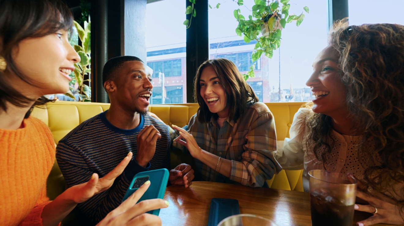 En una cafetería, cuatro amigos ríen mientras uno sostiene un teléfono.