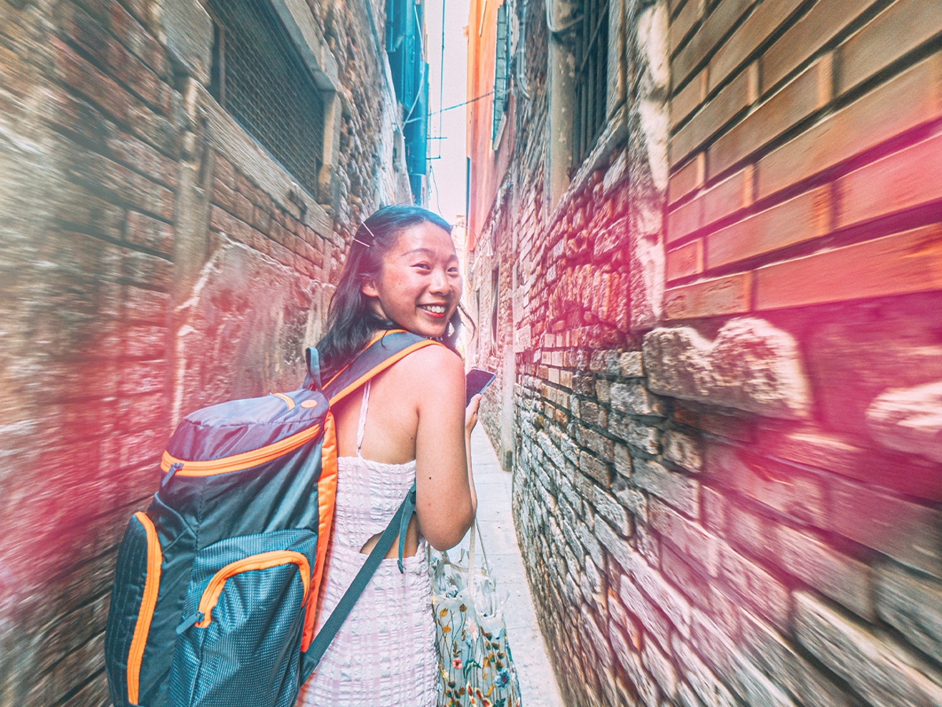 Una joven sonriente caminando por un callejón de ladrillos con una mochila de viaje azul.