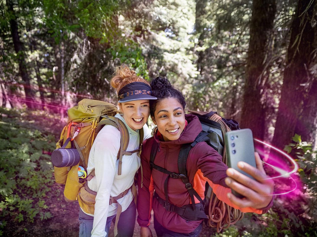Dos mujeres, vestidas con chaquetas y mochilas de acampada, sonríen juntas para tomarse un selfie en un sendero boscoso.
