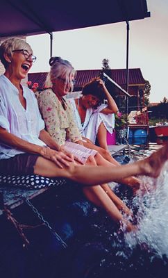 Tres mujeres están sentadas en un muelle y ríen mientras chapotean con los pies en el agua al atardecer.