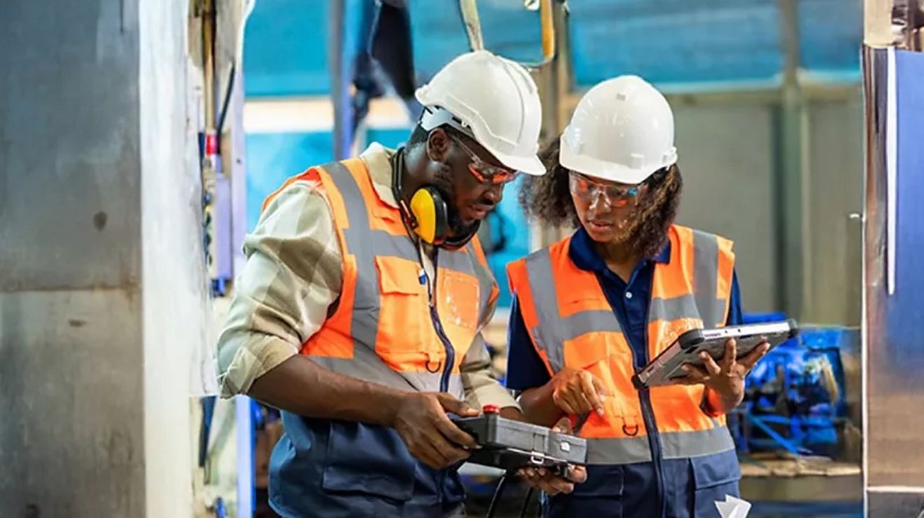 Dos ingenieros mecánicos con casco utilizando equipos conectados de T-Mobile para solucionar problemas con maquinaria industrial.