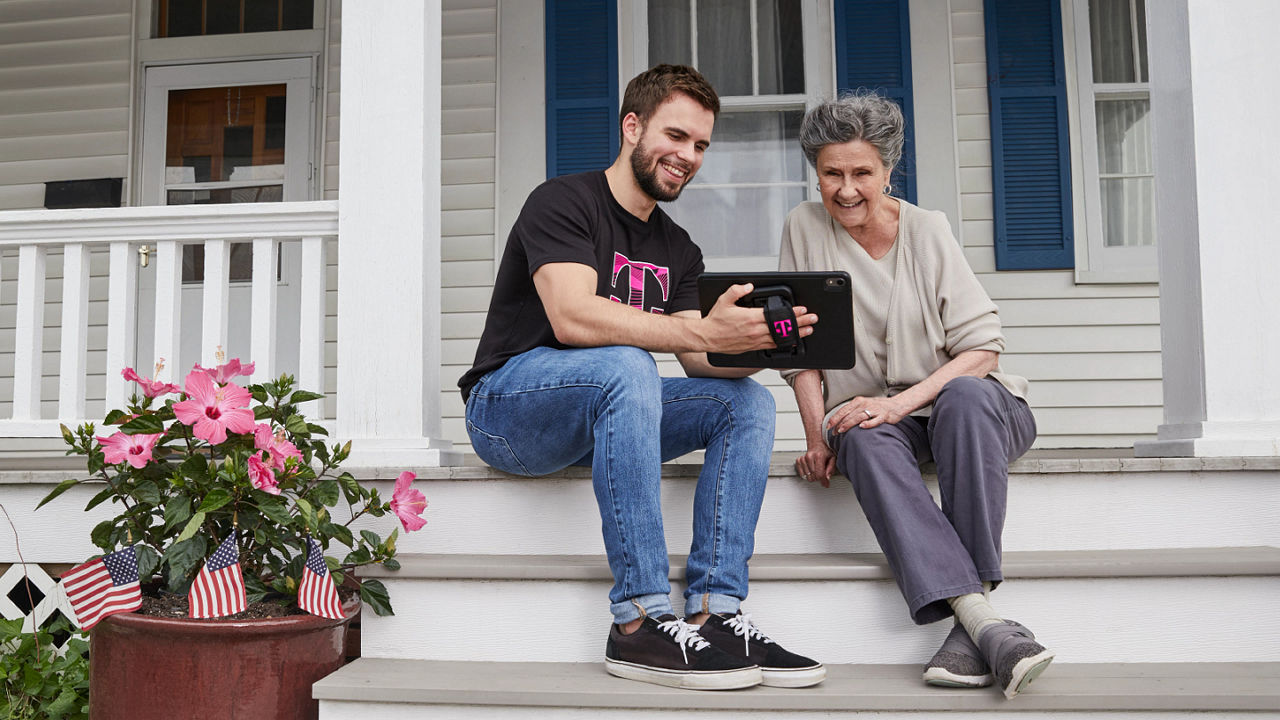Hombre joven y mujer mayor sentados en los escalones delate de una casa mirando una tablet