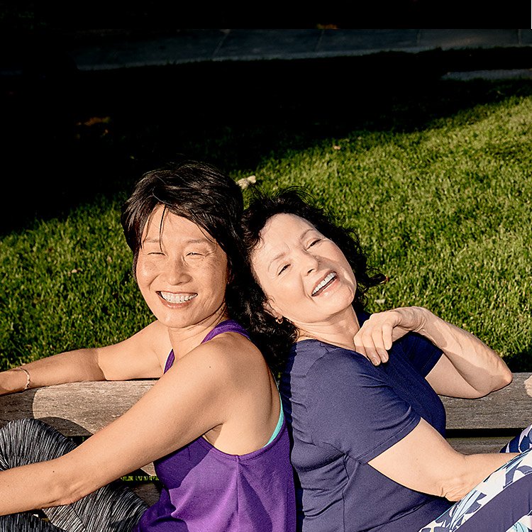 Dos mujeres sonriendo y mirando hacia arriba mientras están espalda con espalda sobre el césped