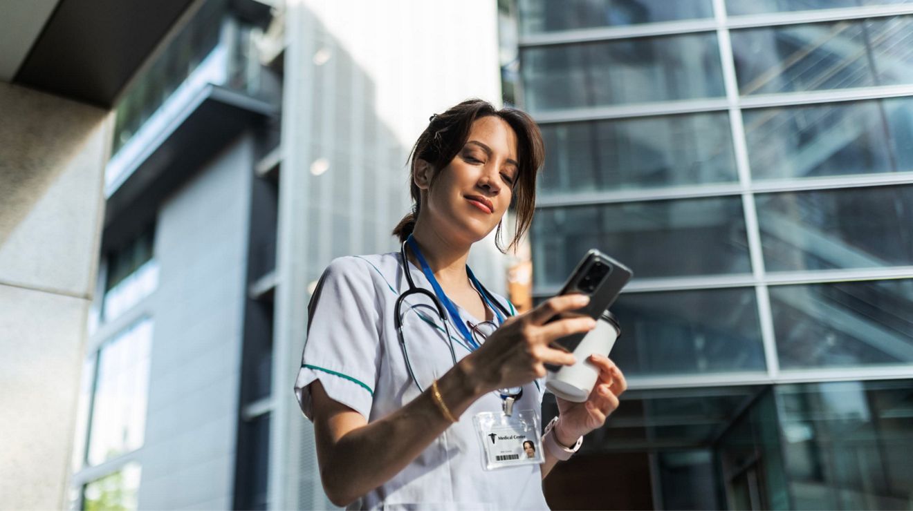Una médica revisando las actualizaciones más recientes en un teléfono en el exterior de un hospital.