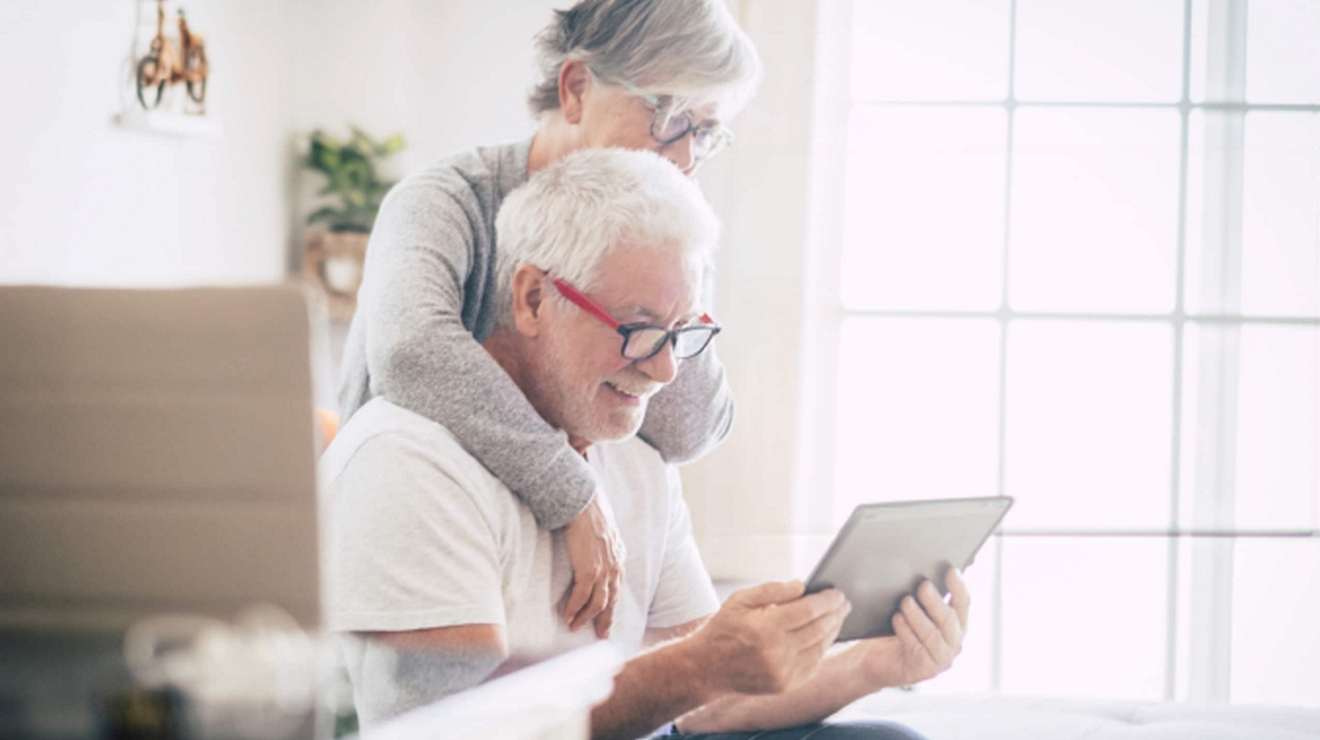 Una pareja de personas mayores se abraza mientras usan una tablet para chatear por video con su familia desde su oficina en casa.