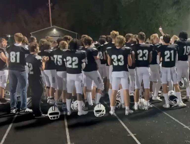 Jugadores de fútbol americano de Port Washington, vestidos con uniformes azul marino y blanco, reunidos en grupo a un lado del campo.