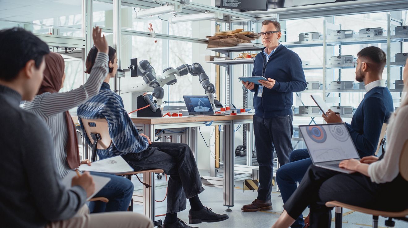 Clase de robótica en la universidad: profesor explica ingeniería a estudiantes utilizando un brazo robótico. Grupo diverso de jóvenes ingenieros programan una mano robótica. Concepto de educación superior en informática.