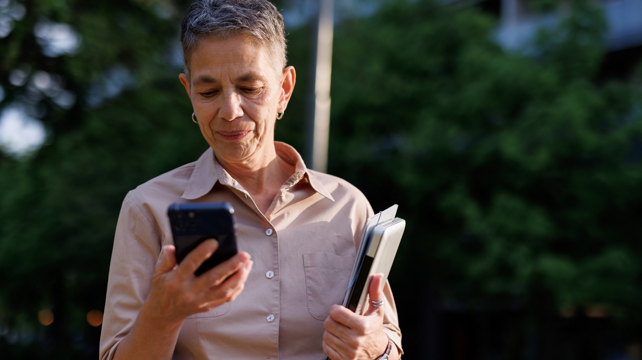 Una profesora mayor sonríe mientras usa su teléfono y sostiene su laptop