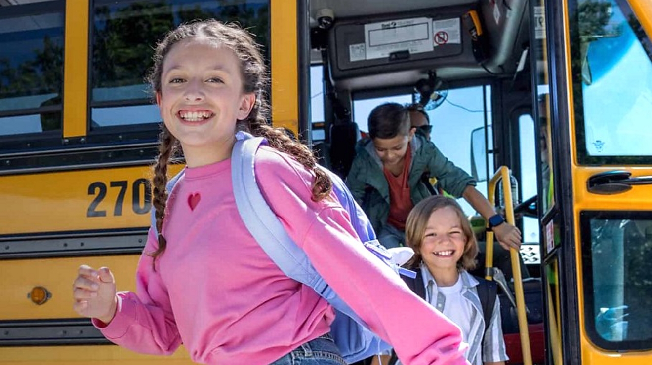 Niña con camiseta rosa sonríe al bajar del autobús escolar.